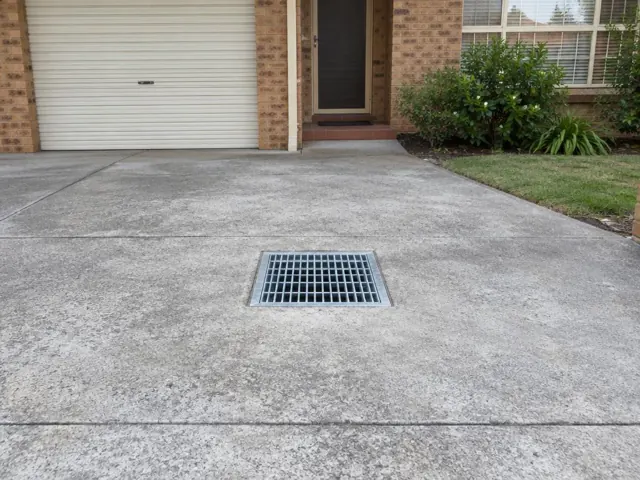 Square outdoor drain grate set in a concrete driveway beside a brick house in Northmead
